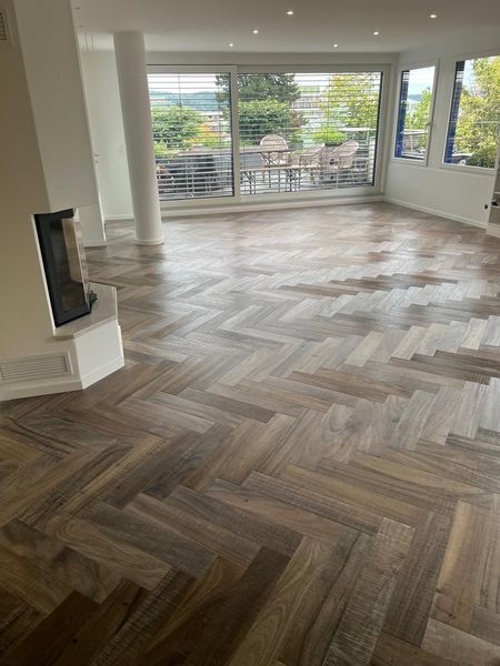 Ein helles Wohnzimmer besticht durch edle, braune Holzdielen im Fischgrätmuster und grosse Fenster mit Blick auf eine Terrasse.