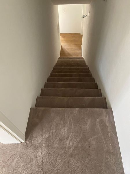 Blick aus der Vogelperspektive auf eine schmale, mit braunem Teppich ausgelegte Innentreppe, die zu einem hellen, holzbedeckten Bereich führt.