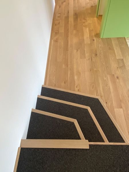 Treppe mit dunkelgrauen, rutschfesten Stufen und hellen Holzkanten, die von einem hellen Holzboden nach unten führt.