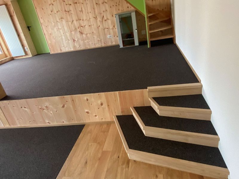 Eine kleine Holztreppe mit dunkel geteerten Stufen führt zu einer erhöhten Plattform mit dunklem Teppichboden.