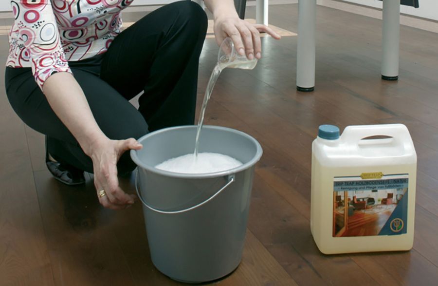 Eine Person giesst Reinigungsmittel in einen grauen Eimer mit Seifenwasser, der auf einem Holzboden neben einem Gallonenkanister steht.