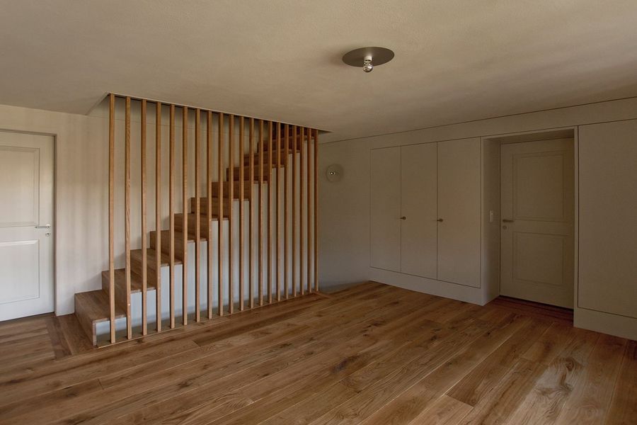Eine Holztreppe mit vertikalen Lattengeländer in einem minimalistisch eingerichteten Raum mit hellen Holzböden und grauen Einbauschränken.