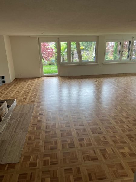 Ein geräumiges, leeres Zimmer mit hellem Parkettboden, weissen Wänden und grossen Fenstern mit Blick auf einen Garten.