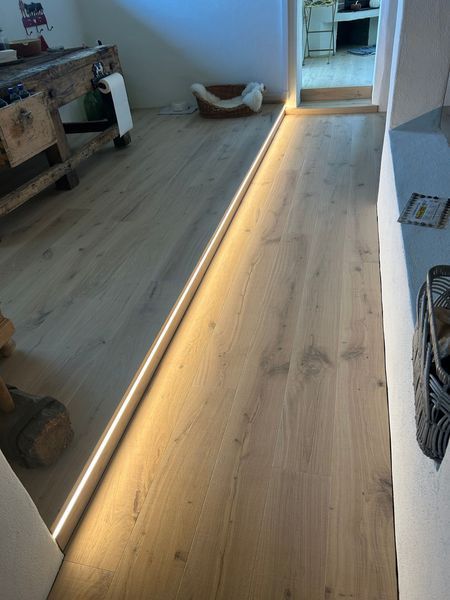 Entlang der Kante eines erhöhten Holzbodens verläuft ein beleuchteter LED-Streifen, der den Übergang zwischen zwei Wohnbereichen erhellt.