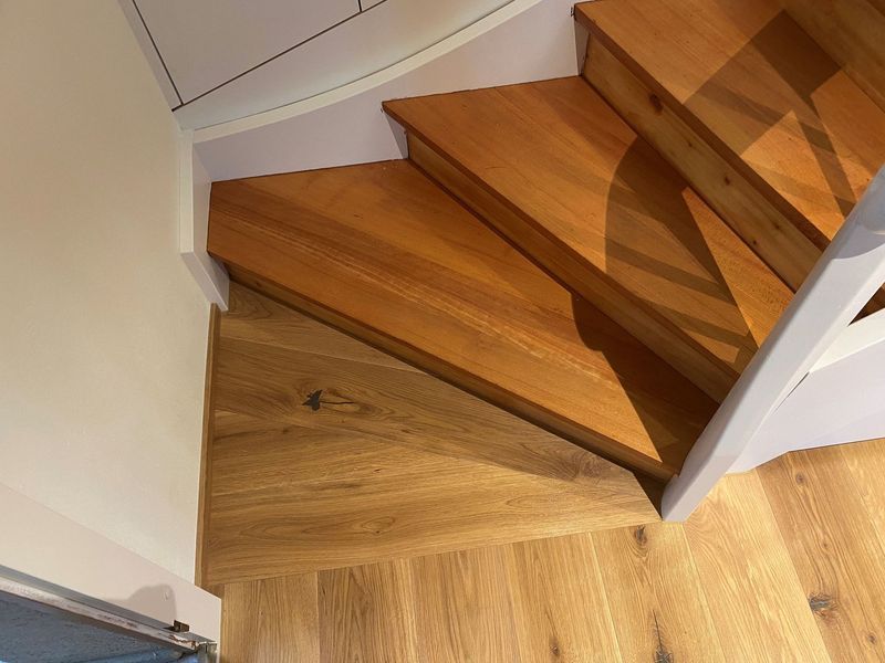 Eine Holztreppe mit keilförmiger unterster Stufe, die in einen hellen Eichenholzboden übergeht, in einem hell erleuchteten Innenraum.