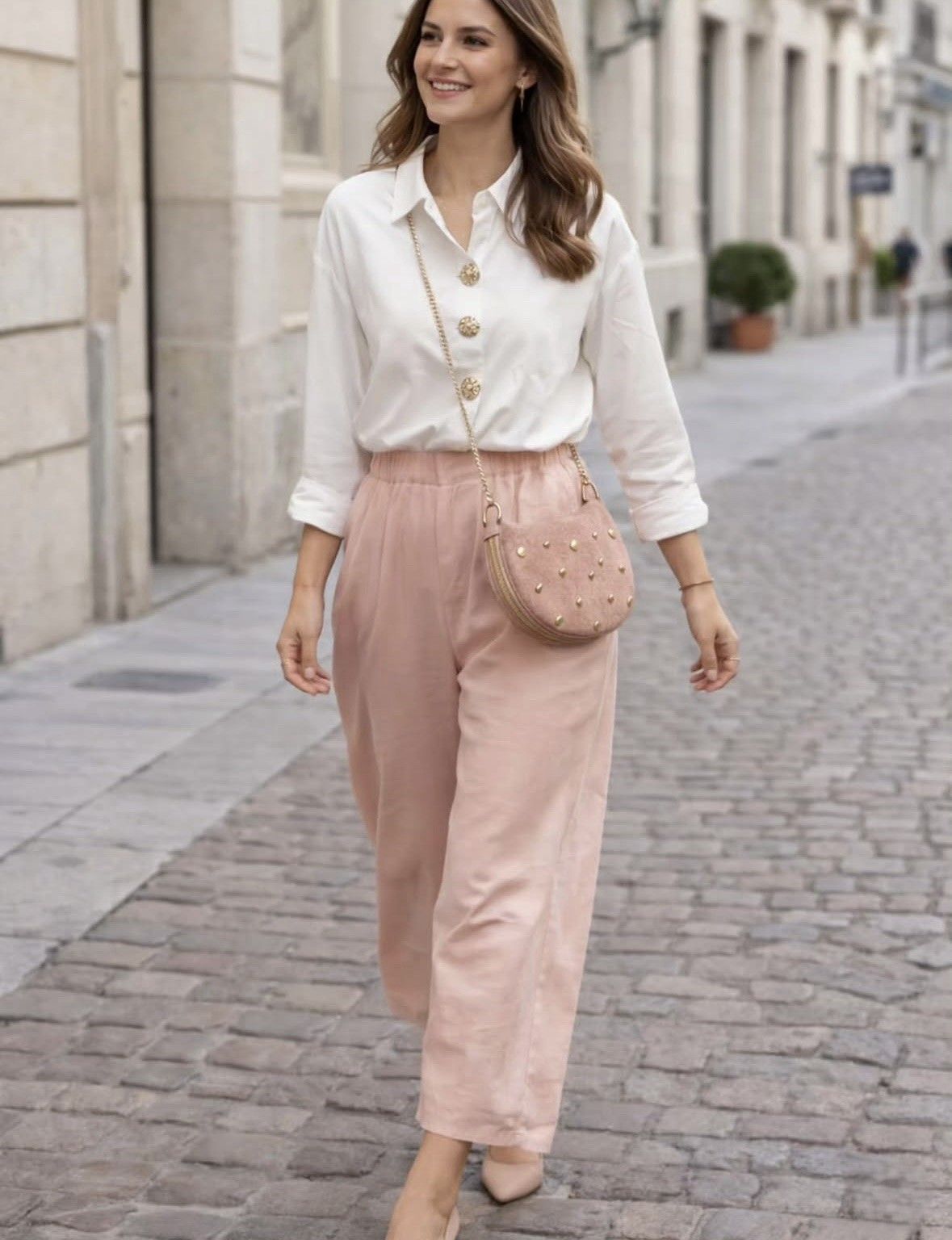 Mujer caminando por una calle empedrada, vestida con una blusa blanca y pantalones rosas con un bolso a juego.
