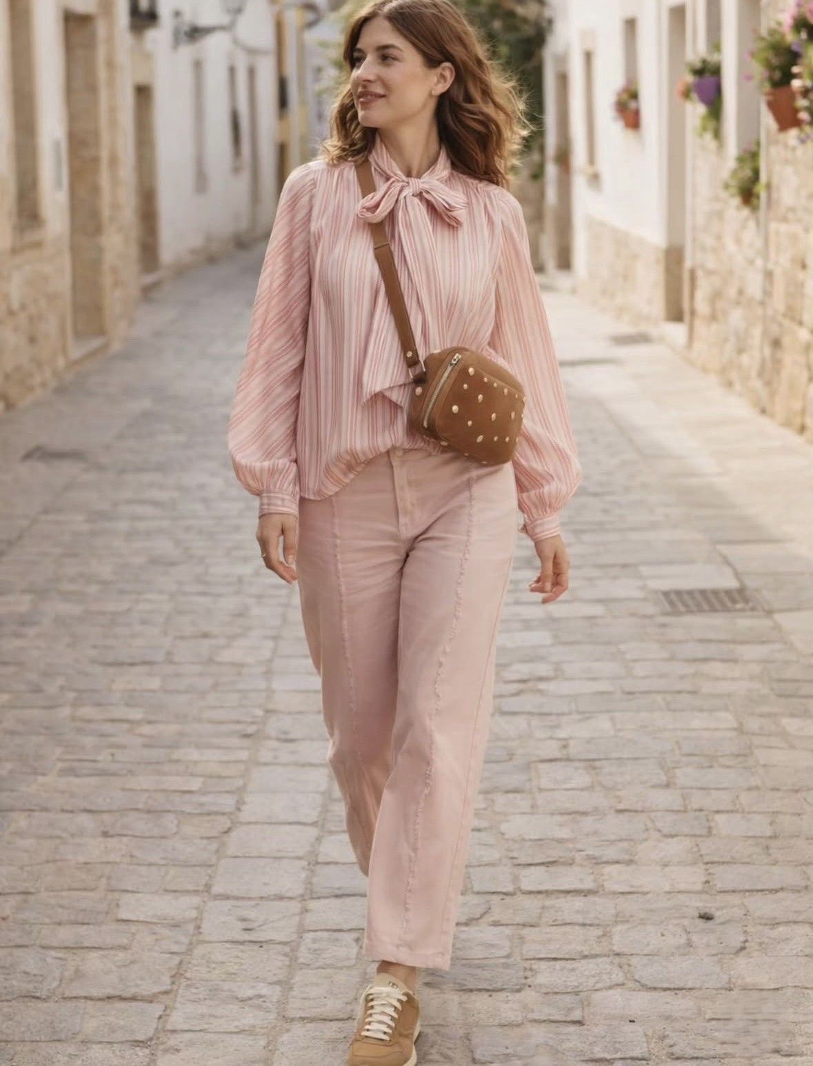 Mujer caminando por una calle empedrada con blusa y pantalones rosas, y un bolso cruzado marrón.
