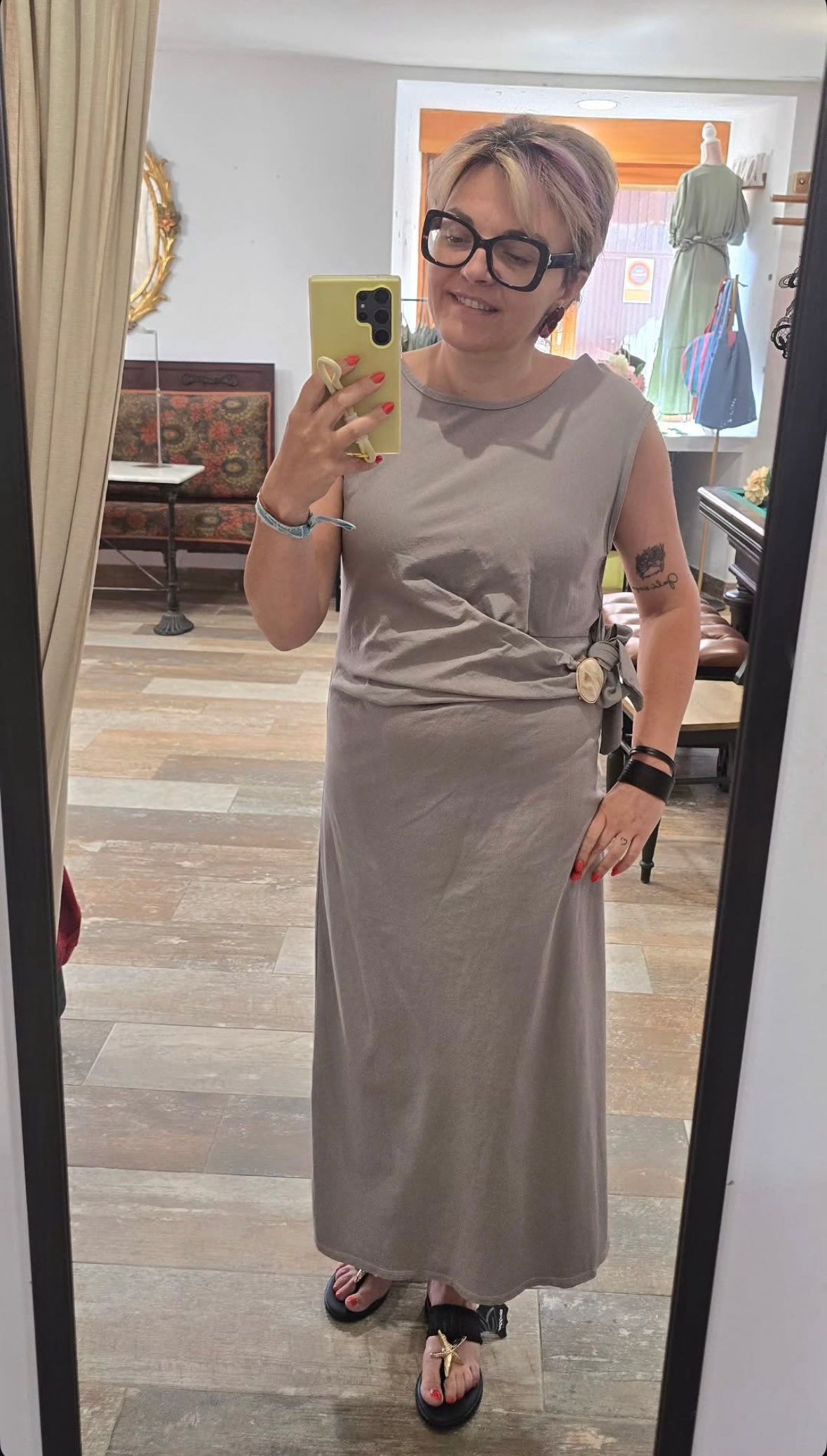 Selfie frente al espejo de una persona con un vestido gris, sosteniendo un teléfono en una habitación luminosa.