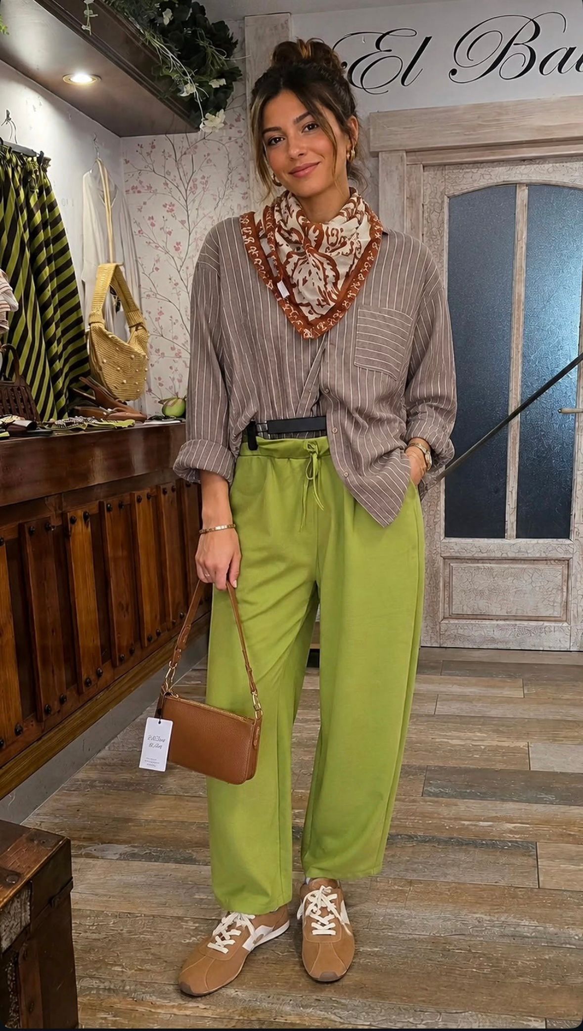 Mujer con blusa estampada y pantalones verde lima posando en interiores con un bolso marrón y un collar llamativo.