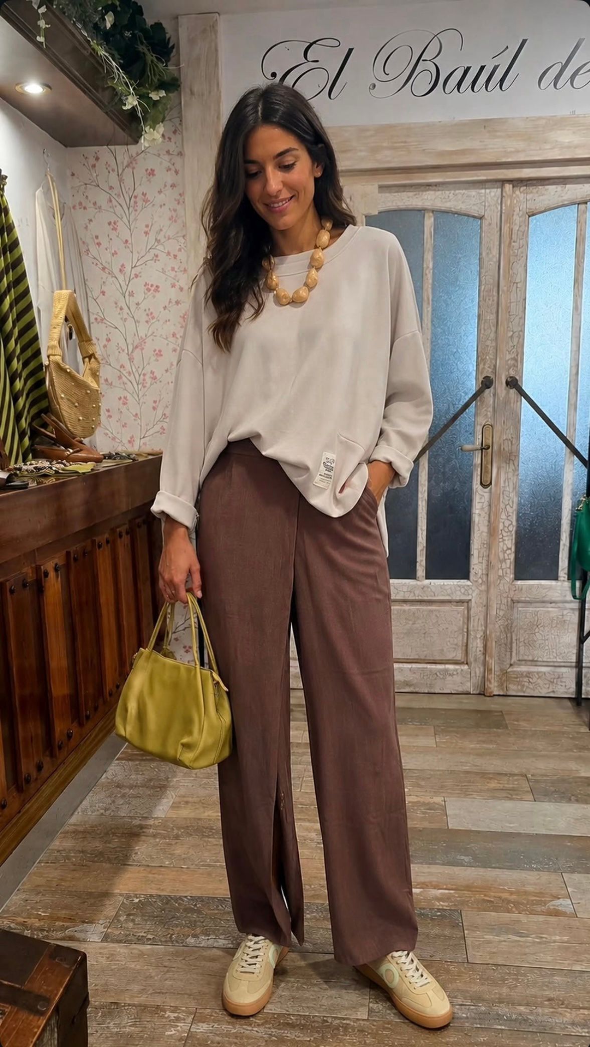 Mujer de pie en el interior de una habitación rústica, vestida con una blusa blanca y pantalones marrones