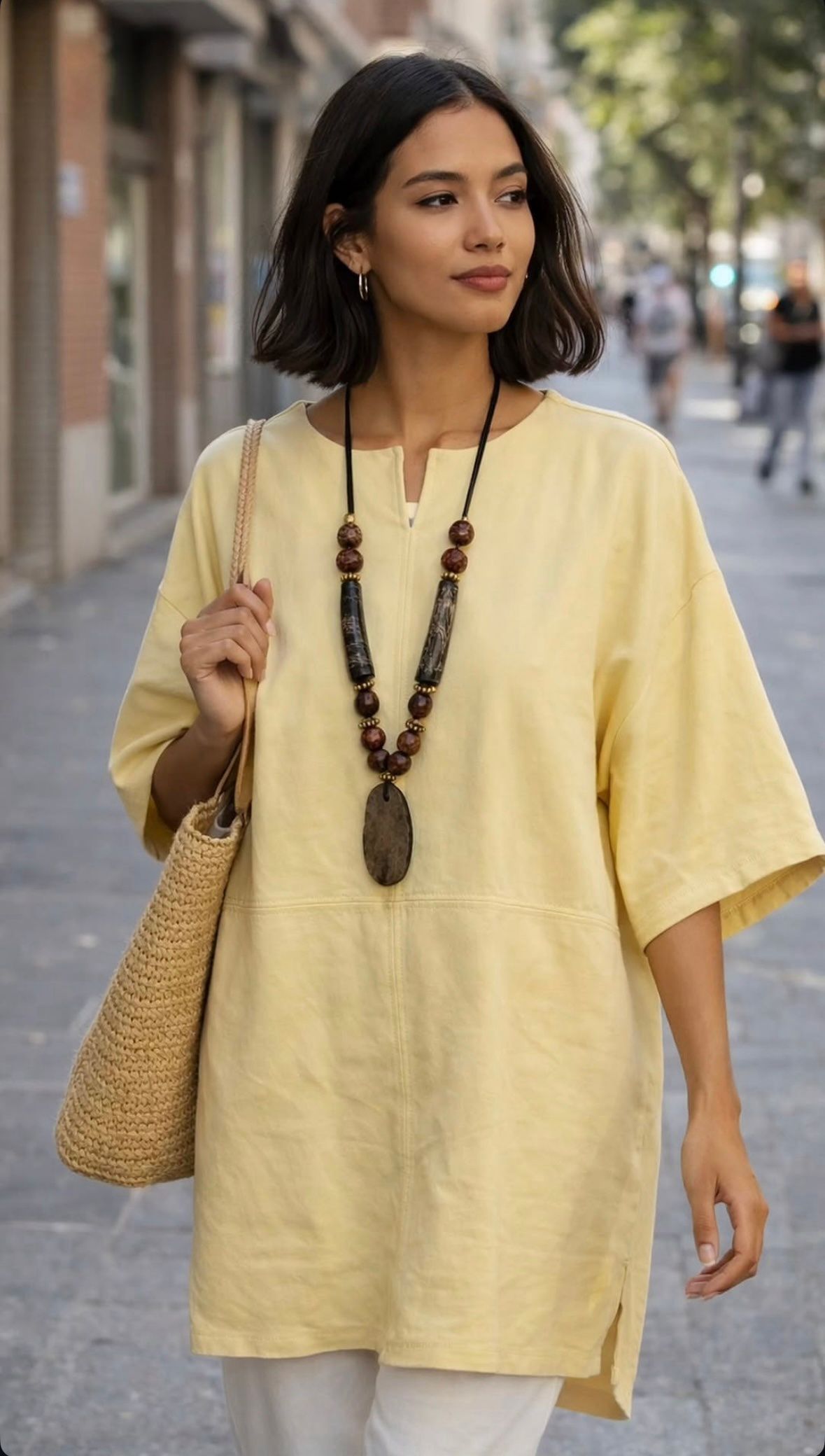 Mujer con túnica amarilla holgada, collar de cuentas y bolso de paja caminando al aire libre por una calle de la ciudad.