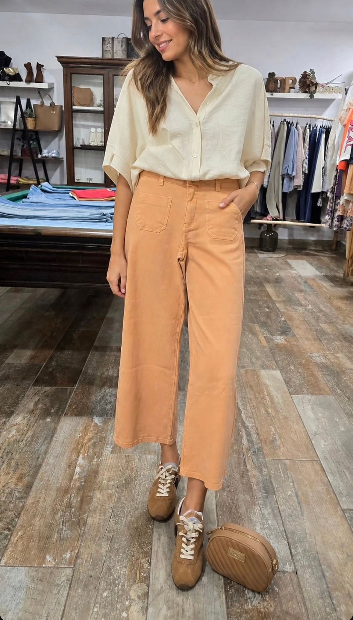 Mujer con blusa color crema y pantalones color canela de pie en una tienda de ropa, con una bolsa de mimbre en el suelo.