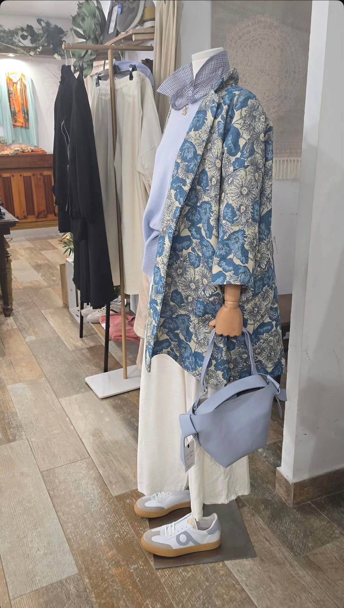 Maniquí con chaqueta azul estampada de flores y pantalones blancos junto a un perchero en una boutique.