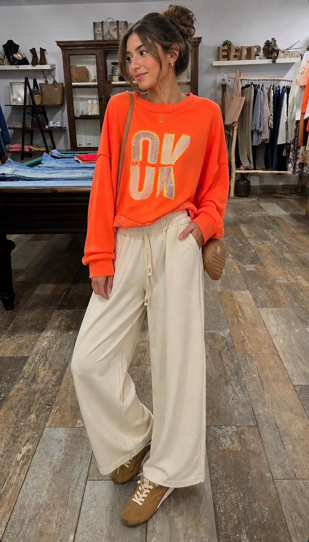 Mujer posando con un suéter naranja brillante y pantalones anchos de color beige en una tienda de ropa.