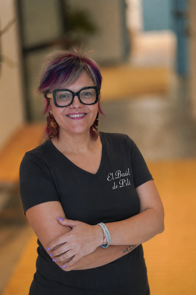 Persona sonriente con gafas y pelo morado, de pie con los brazos cruzados en un pasillo.