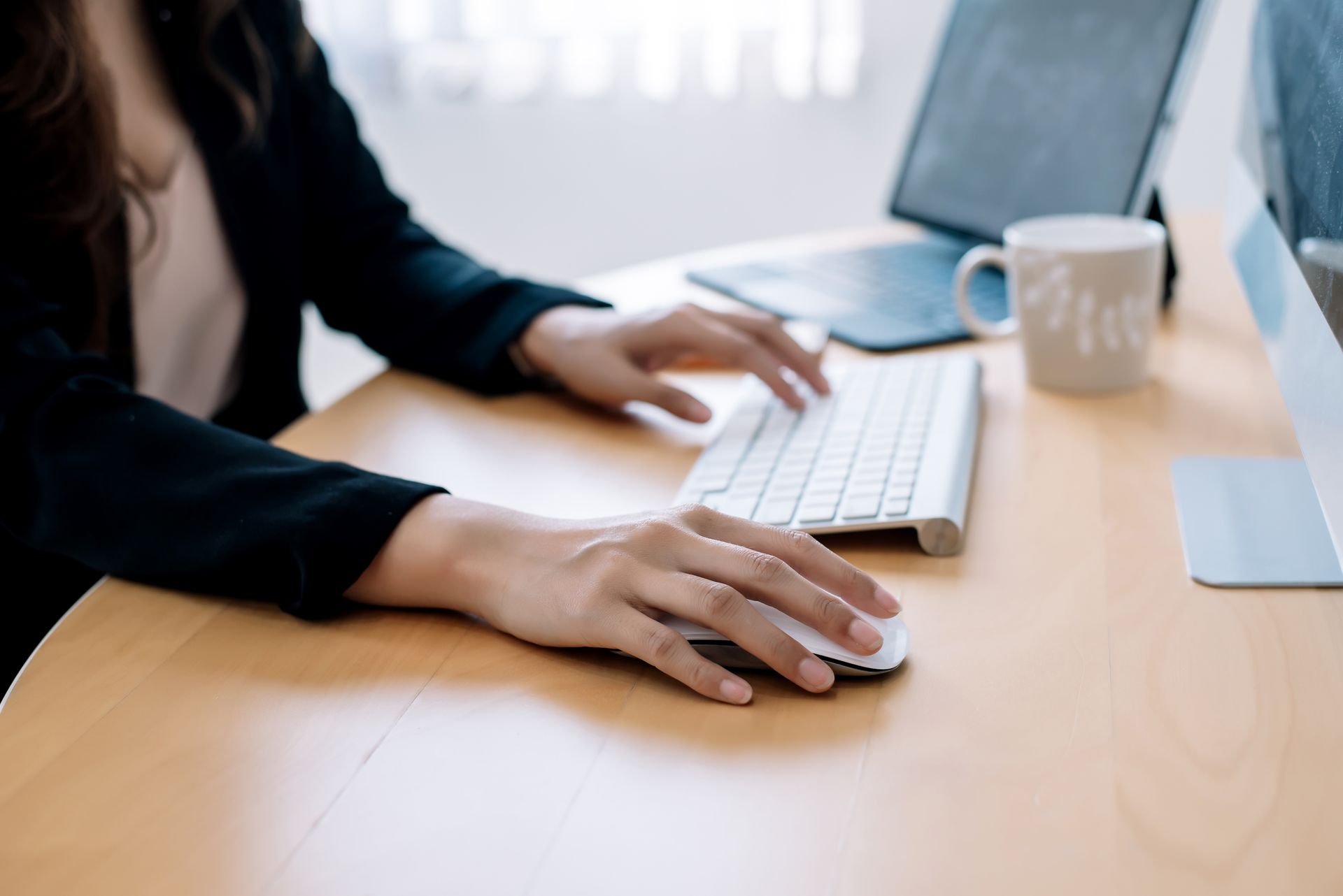 Les mains d'une femme tapant sur un clavier, utilisant une souris à un bureau avec un écran d'ordinateur, un ordinateur portable et une tasse.