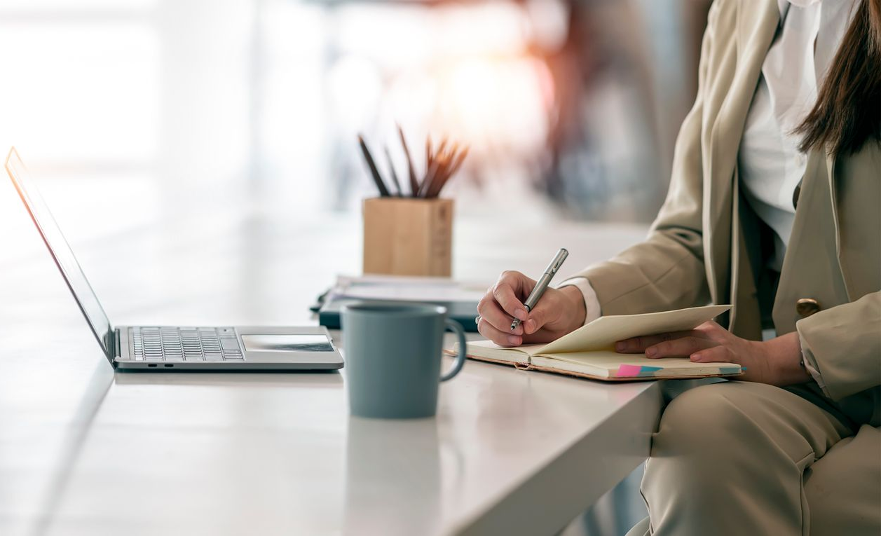 Une femme en tailleur écrit dans un carnet à un bureau où se trouvent un ordinateur portable et une tasse.