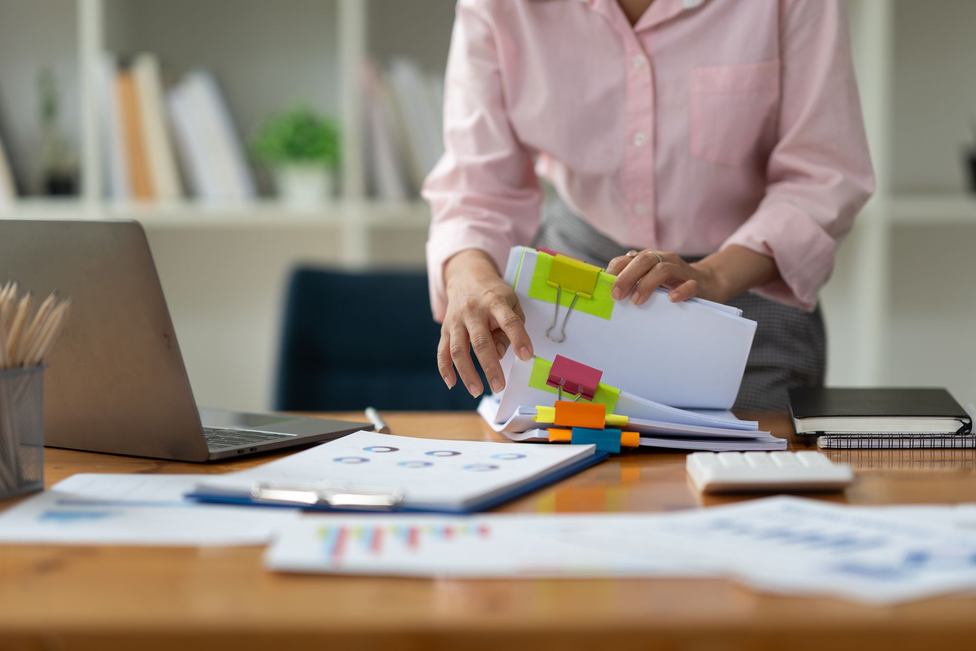 Une femme trie des documents sur un bureau, avec un ordinateur portable, des graphiques, une calculatrice et un carnet, dans un bureau.