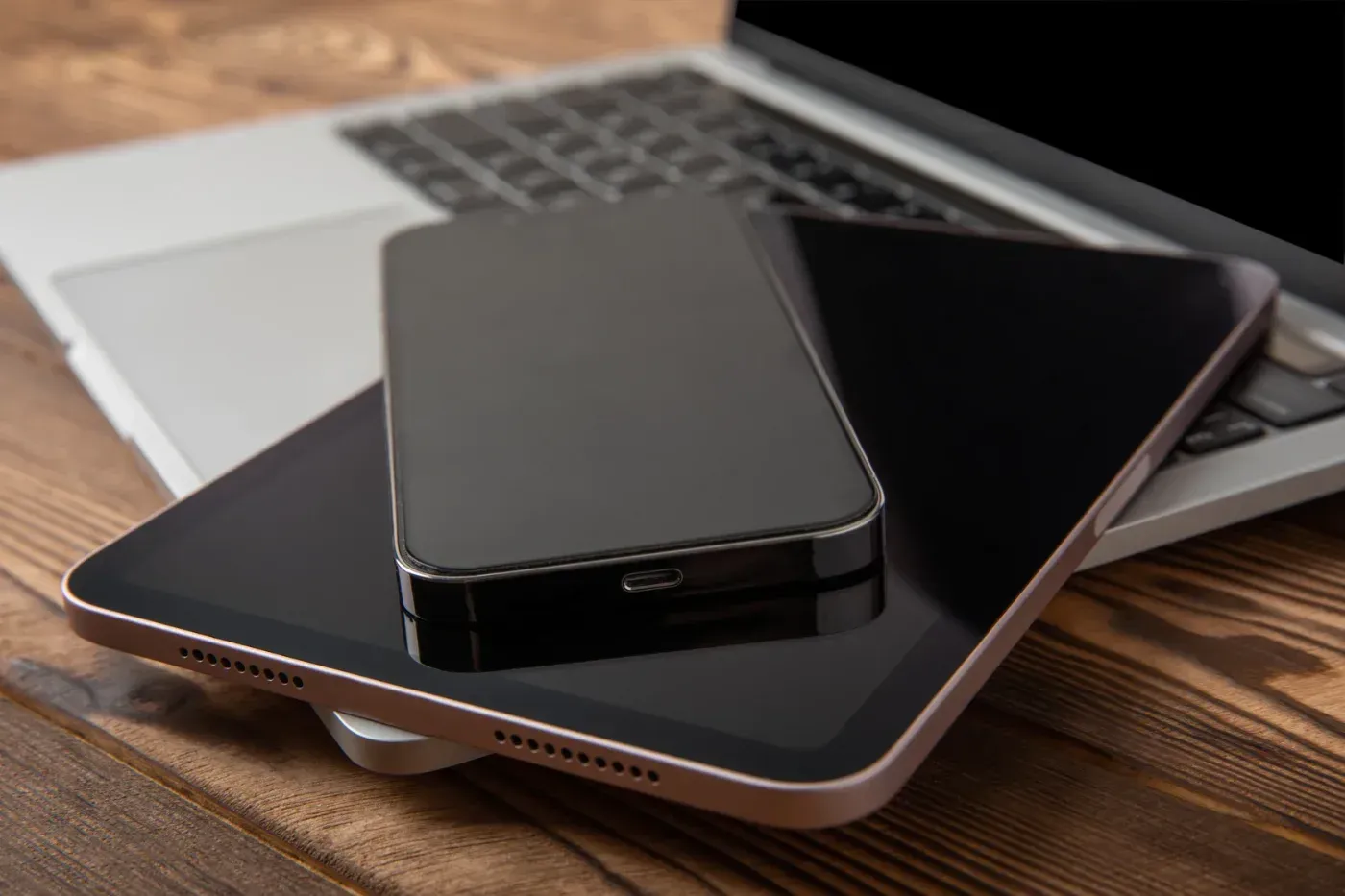 Laptop, Tablet und Smartphone auf einer Holzoberfläche gestapelt. Das Telefon liegt auf dem Tablet.