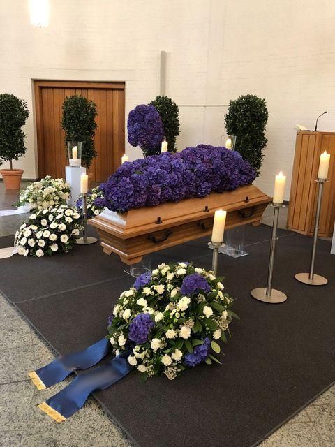 Ein mit lila Blumen bedeckter Sarg bei einer Beerdigung. Weiße und blaue Blumenarrangements, brennende Kerzen.