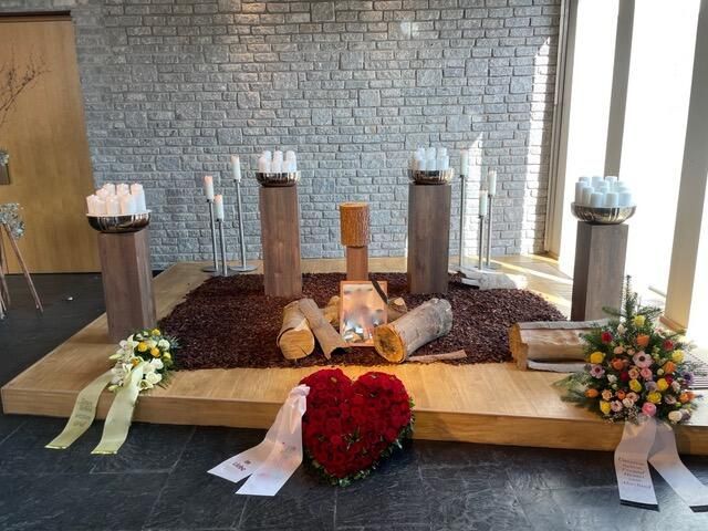 Begräbnisaltar mit Blumen, Kerzen und Holzscheiten. Steinmauer im Hintergrund.