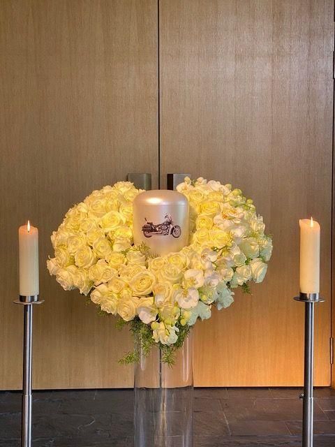 Urne mit Motorradmotiv auf Blumenkranz, flankiert von brennenden Kerzen, in Gedenkumgebung.