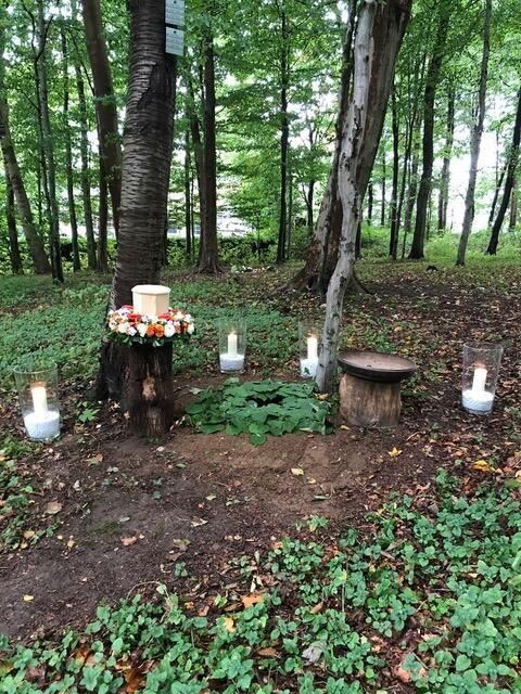 Grabstätte im Wald: Urne auf einem Baumstumpf, Blumenkranz, Kerzen, kreisförmige Bestattung, umgeben von Bäumen.