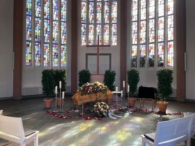 Trauerfeier in einer Kapelle, Sarg mit Blumen, Buntglasfenstern, Kerzen und leeren Stühlen.