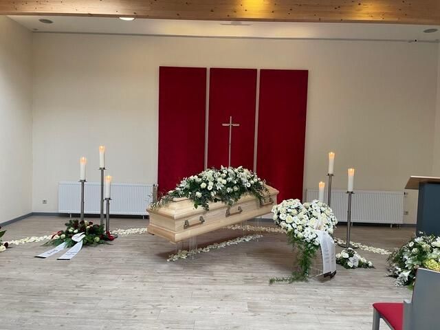 Trauerzeremonie: Sarg mit weißen Blumen, Kerzen, rotem Hintergrund und Kreuz.