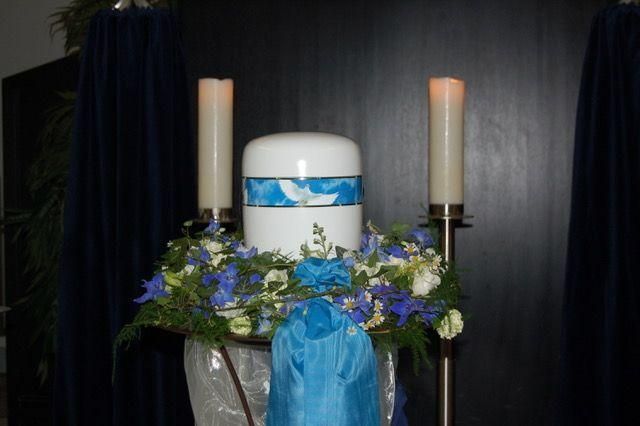 Weiß-blaue Urne mit Blumen, Kerzen und einem drapierten blauen Stoff.