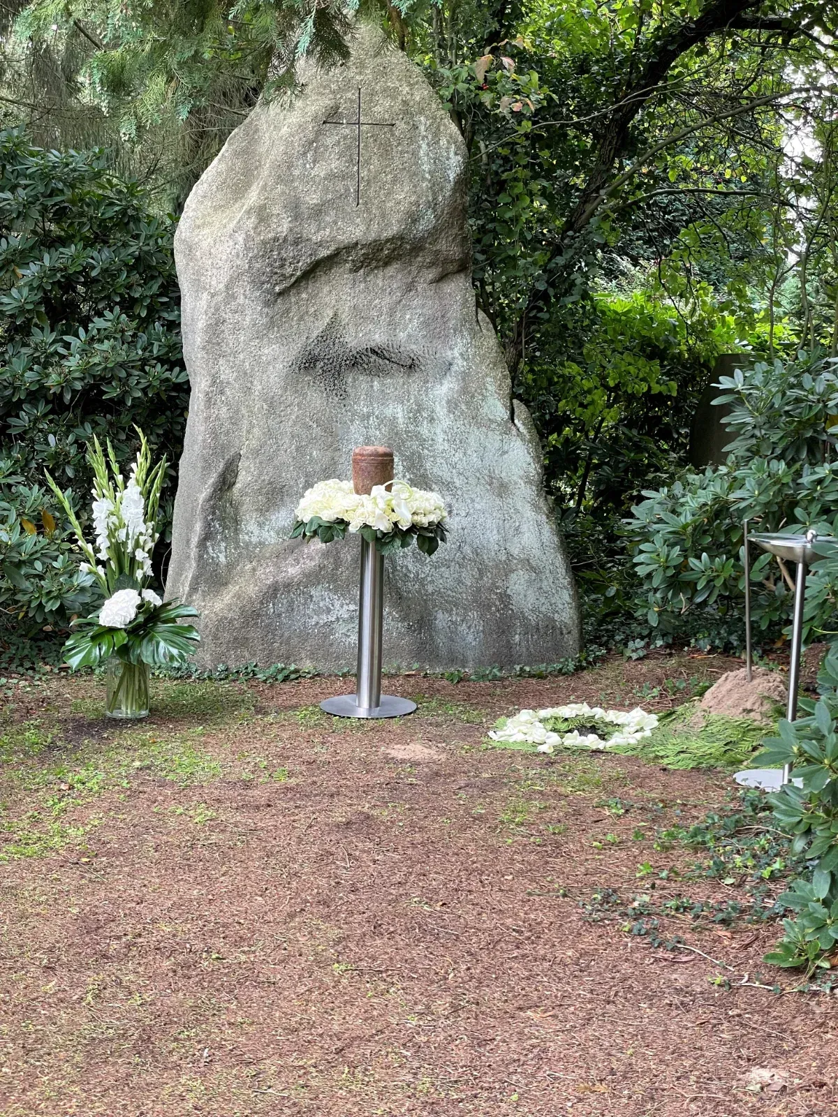 Ein Denkmal mit einem großen Stein, Blumen und einer Urne. In den Stein gehauenes Kreuz.