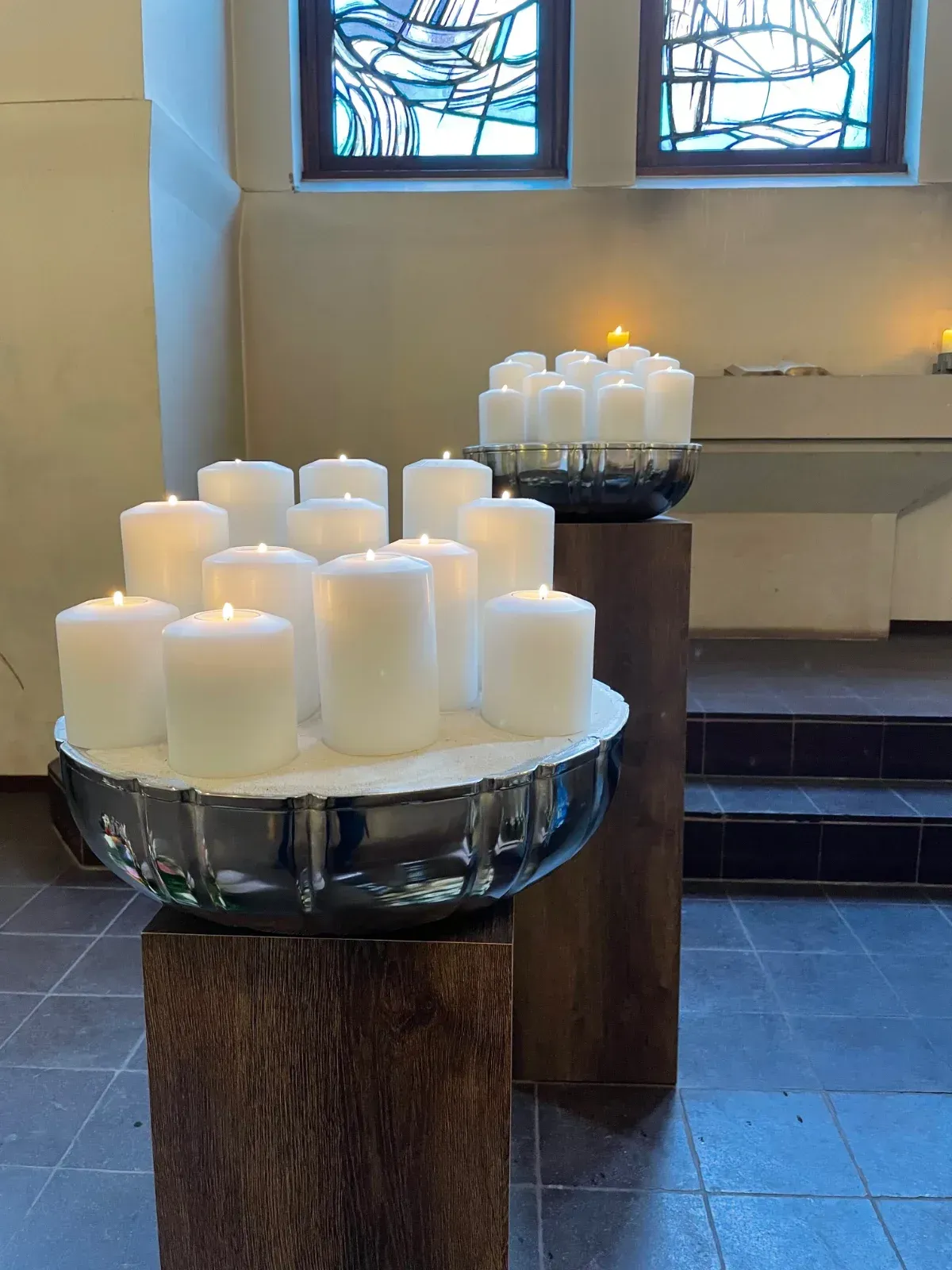 Zwei Sätze weißer Kerzen brennen in einer Kirche und stehen auf Holzsockeln mit einem Buntglasfenster im Hintergrund.