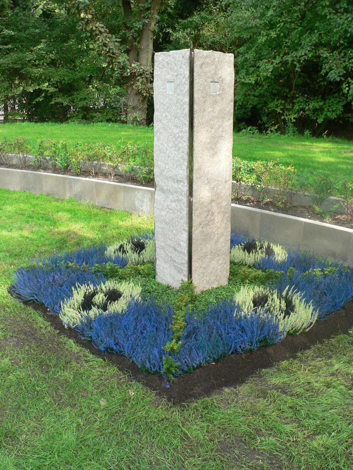 Zwei Steinsäulen in einem Gartenbeet mit blauen und grünen Pflanzen; grüner Grashintergrund.