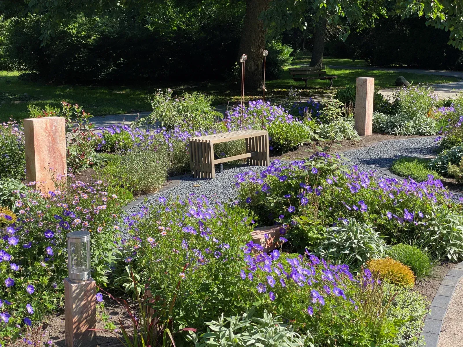Ein Garten mit einer Holzbank, umgeben von blauen Blumen, Steinsäulen und einem Kiesweg.