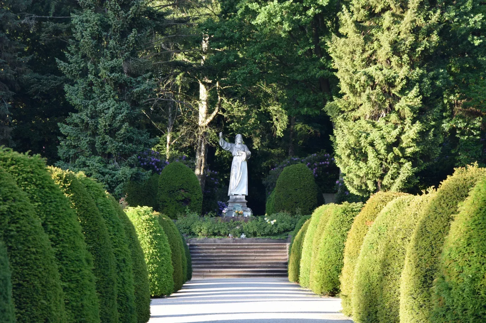 Eine Engelsstatue am Ende eines von gestutzten Büschen und Bäumen gesäumten Pfades.