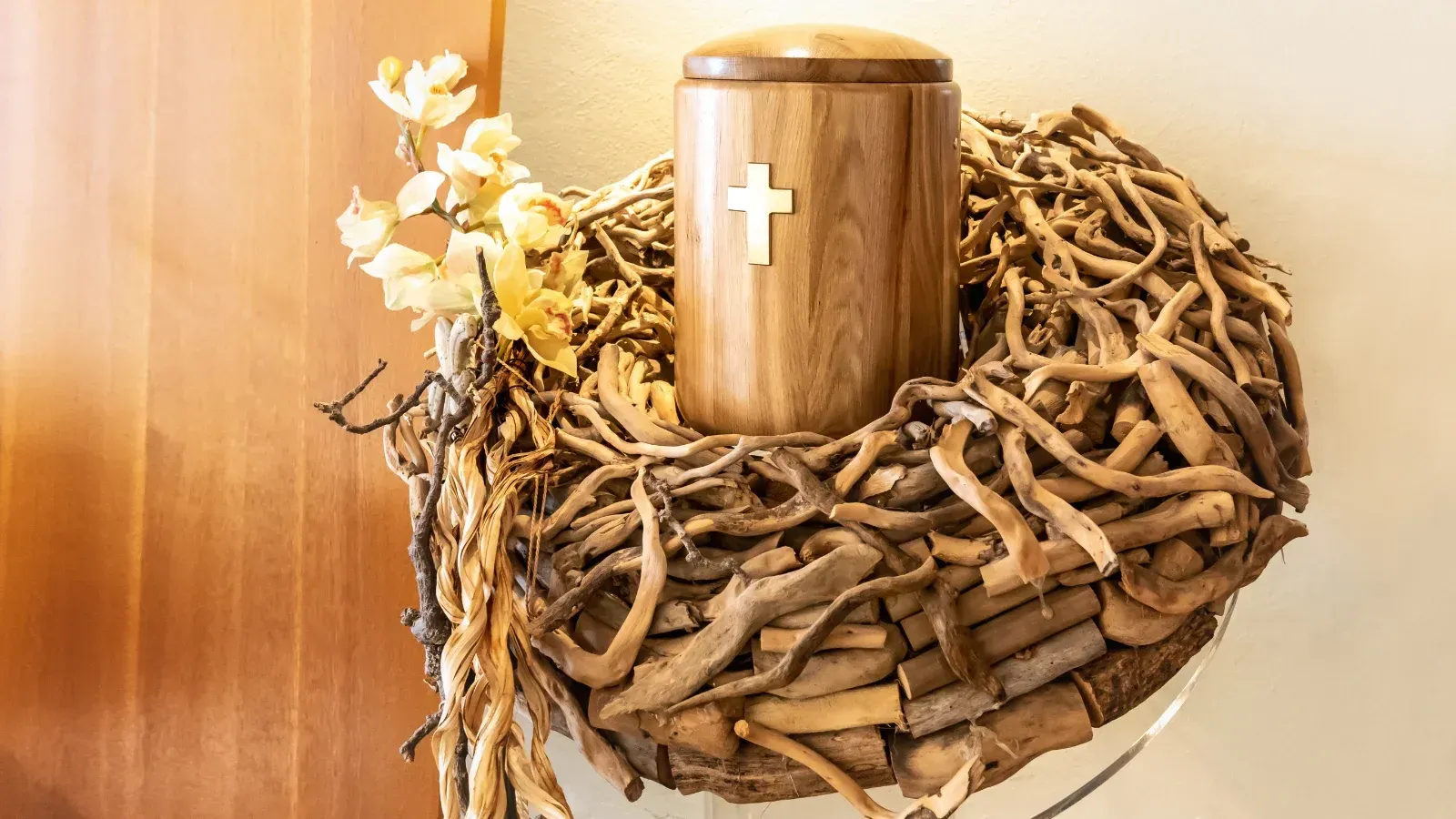 Holzurne mit Kreuz, umgeben von einem Kranz aus Zweigen und Blumen, auf einem Regal.