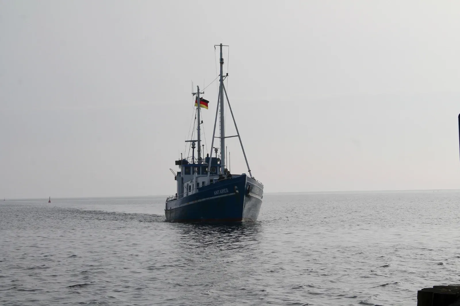 Ein Fischerboot fährt auf ruhigem Wasser unter grauem Himmel, die deutsche Flagge weht aus der Bugperspektive.
