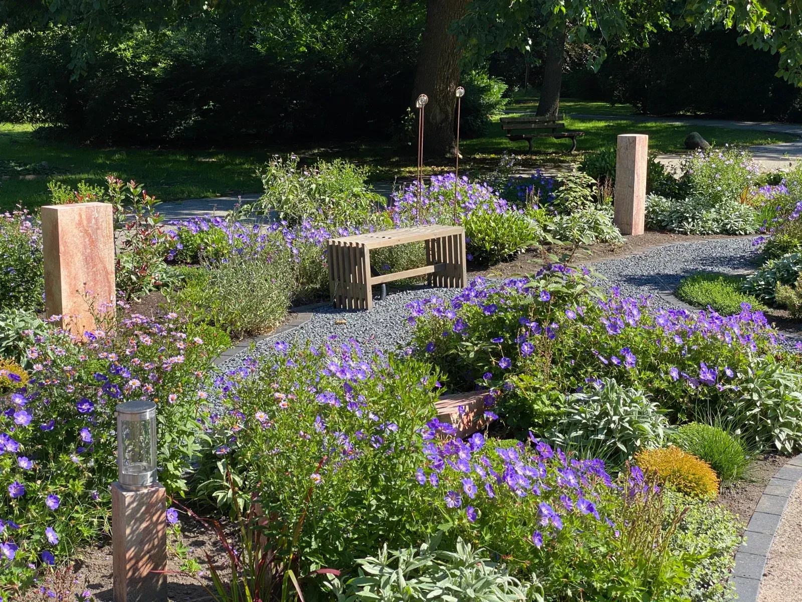 Steinsäulen und eine Bank inmitten eines Gartens mit lila Blumen und viel Grün.