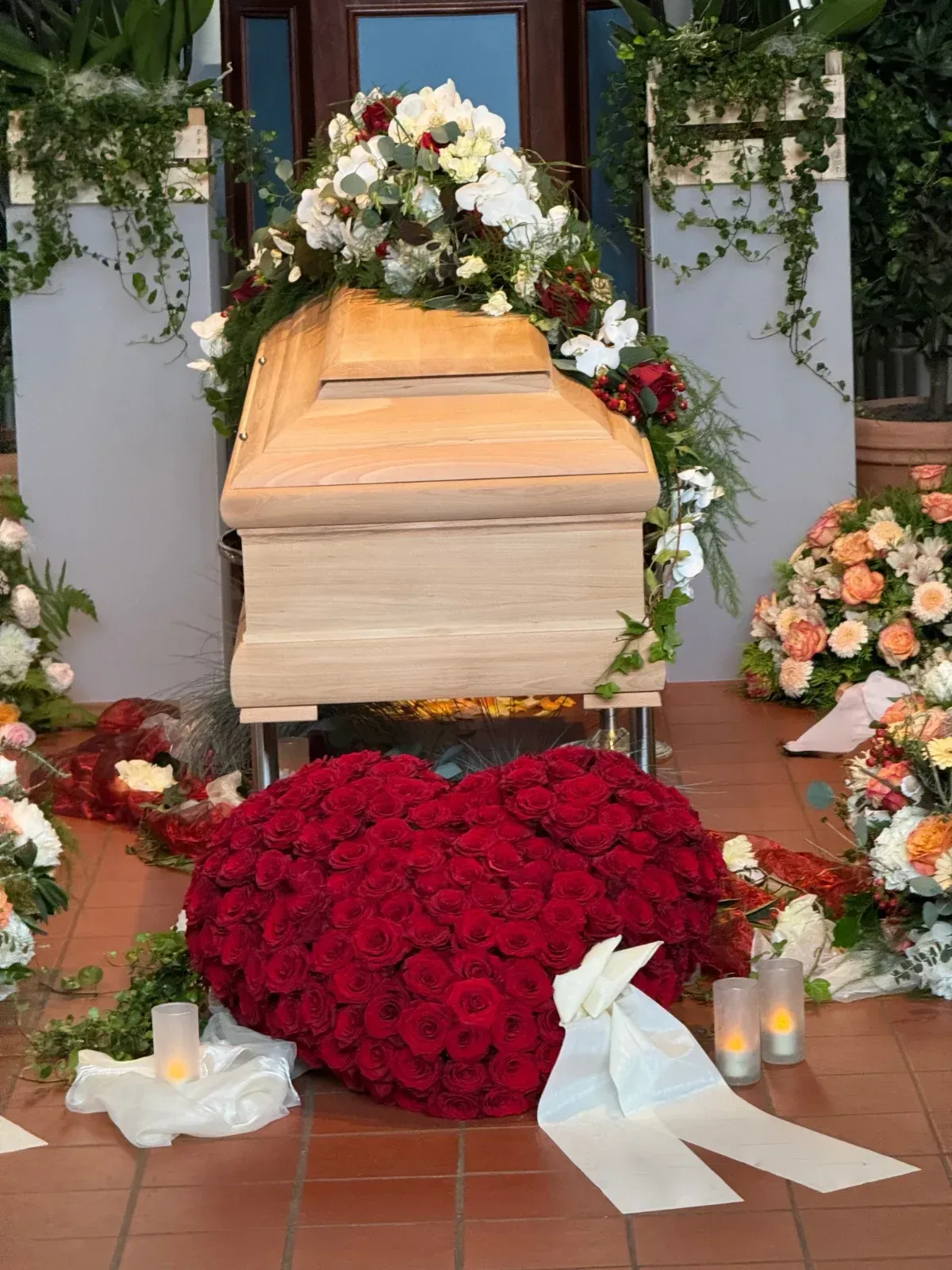 Ein heller Sarg, umgeben von Blumenarrangements, darunter ein rotes Rosenherz.