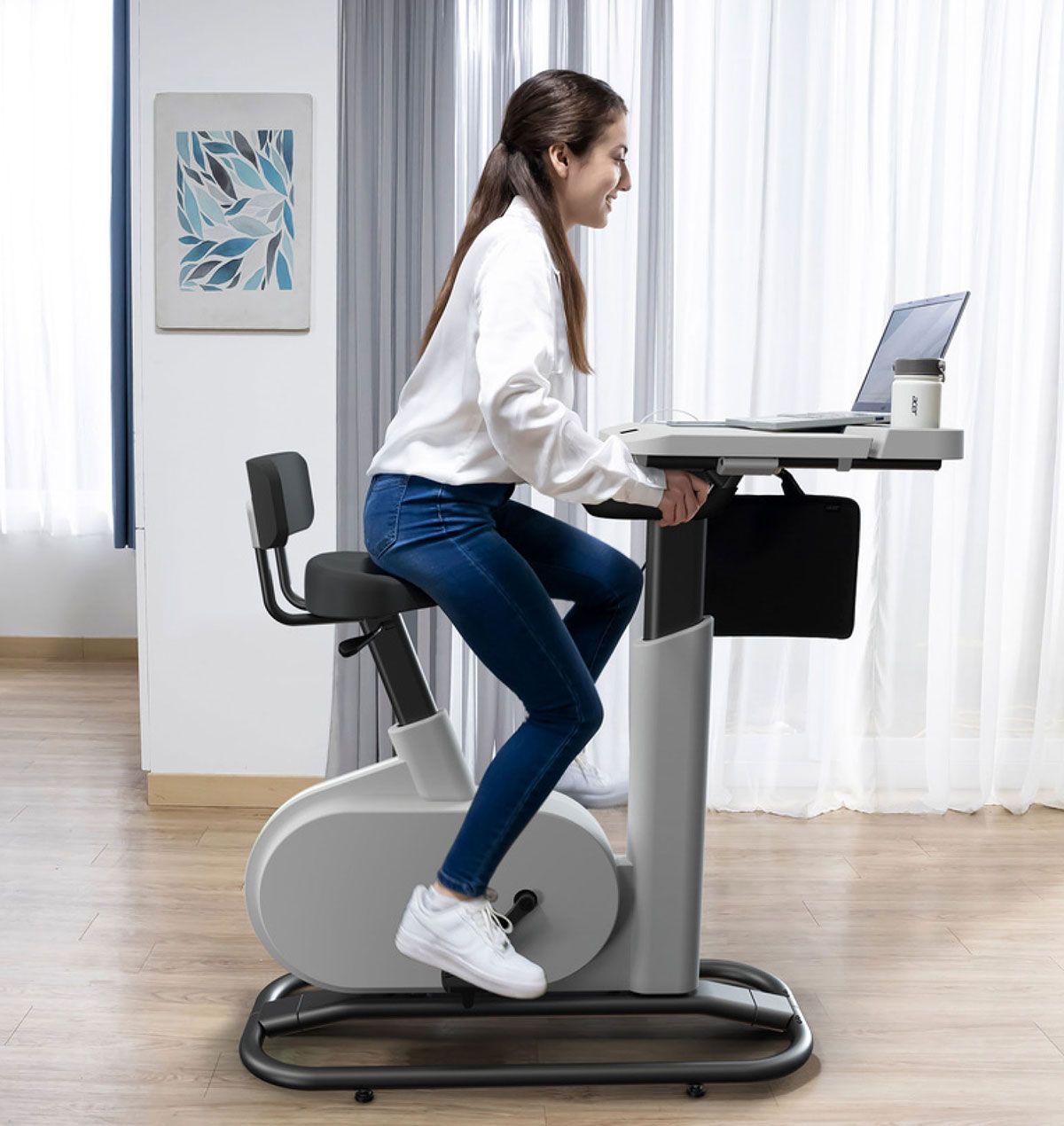 Une femme utilise un bureau pour vélo stationnaire et travaille sur un ordinateur portable. Elle sourit dans une pièce lumineuse.