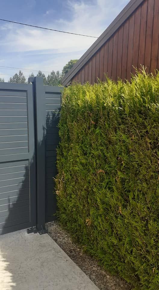 Clôture grise à côté d’une haute haie verte et d’un bâtiment en bois brun, le long d’un chemin en béton.