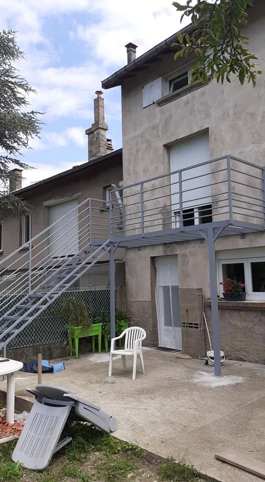 Patio arrière avec une maison grise, un escalier en métal et des chaises en plastique blanc sous un ciel couvert