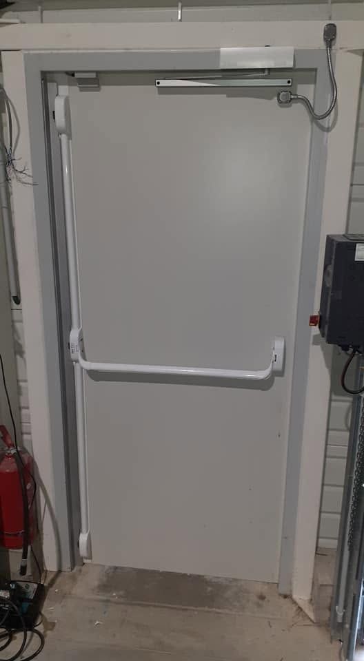 Porte industrielle grise avec barre anti-panique, câblage électrique au-dessus, dans un local technique carrelé