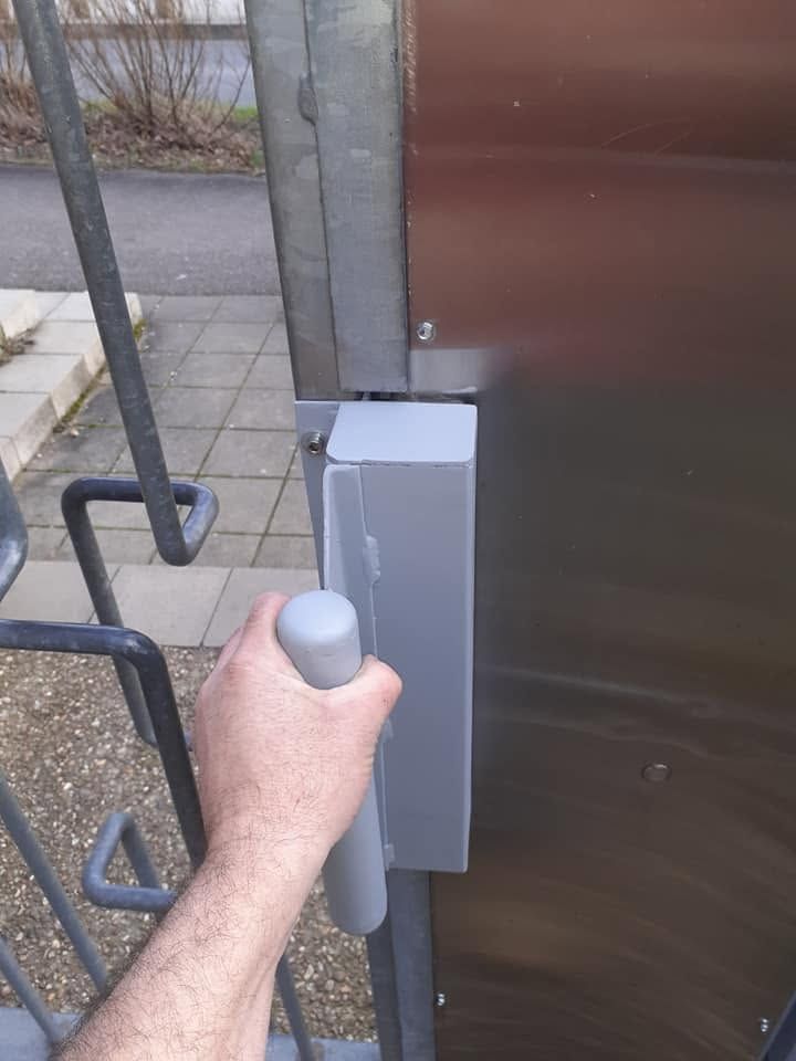 Une main agrippant un loquet gris sur un portail, à côté d’une clôture métallique et d’un bardage brun