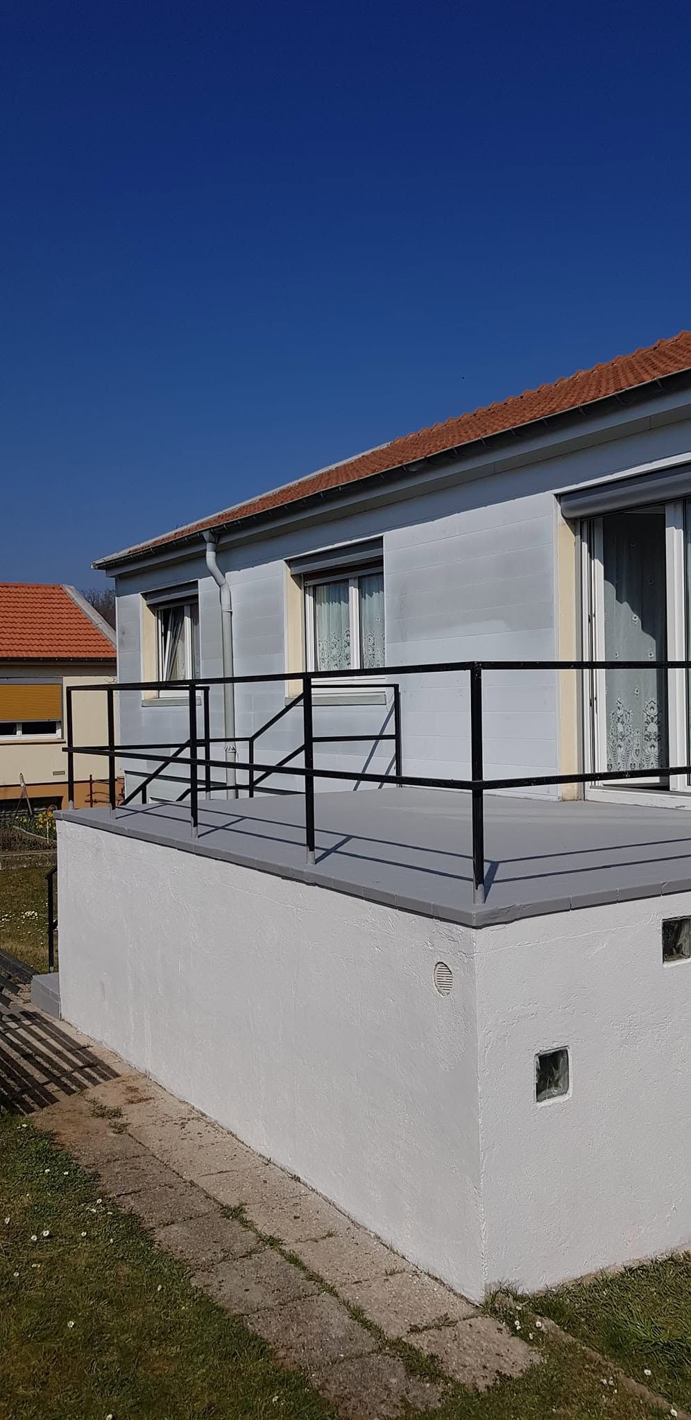 Maison blanche avec un toit rouge, une rampe noire et une terrasse avec une rampe d’accès, sous un ciel bleu dégagé.