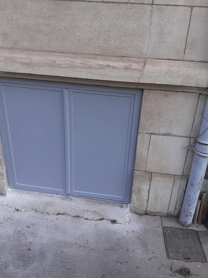 Porte de sous-sol en métal gris intégrée à un mur en béton, à côté d’une descente d’eau pluviale et d’un trottoir.