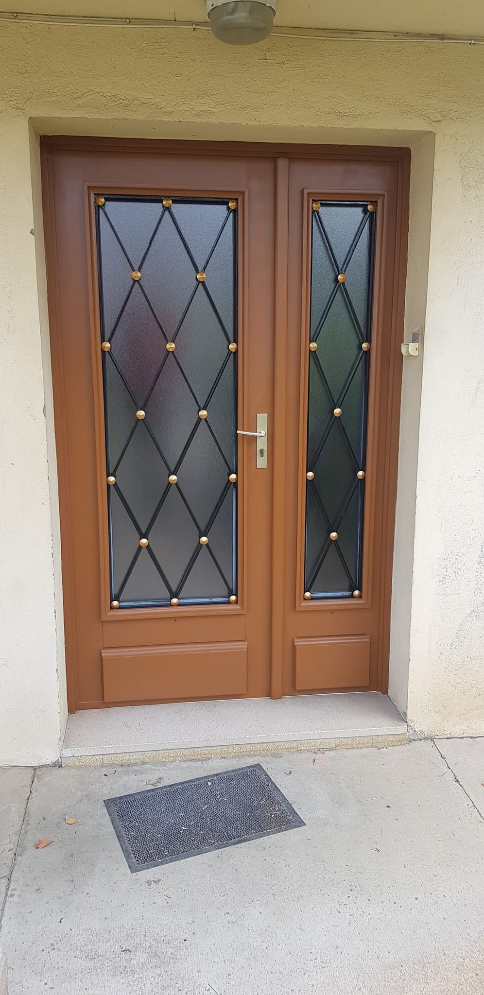 Porte d’entrée double marron avec des panneaux vitrés à motifs losangés et un paillasson devant une entrée en stuc.