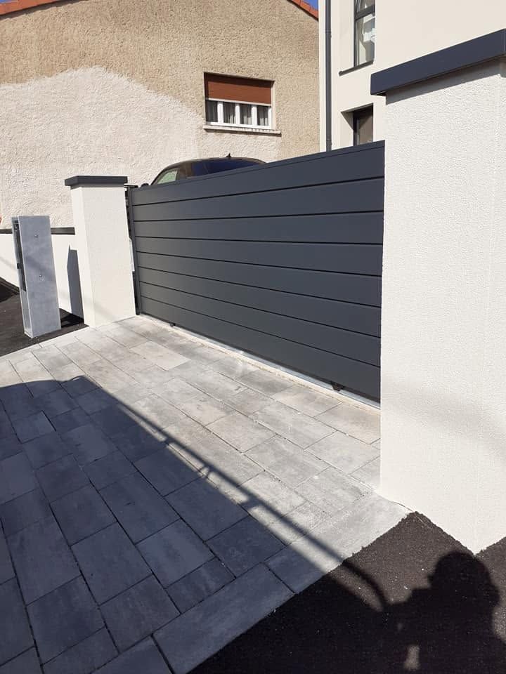 Entrée d’une maison moderne avec un portail coulissant gris foncé, des murs blancs et une allée pavée ensoleillée
