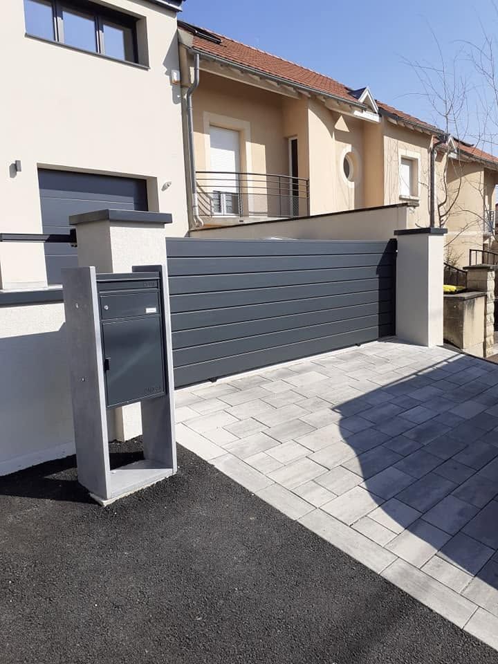 Maison moderne beige avec un portail gris donnant sur une allée pavée, en plein soleil