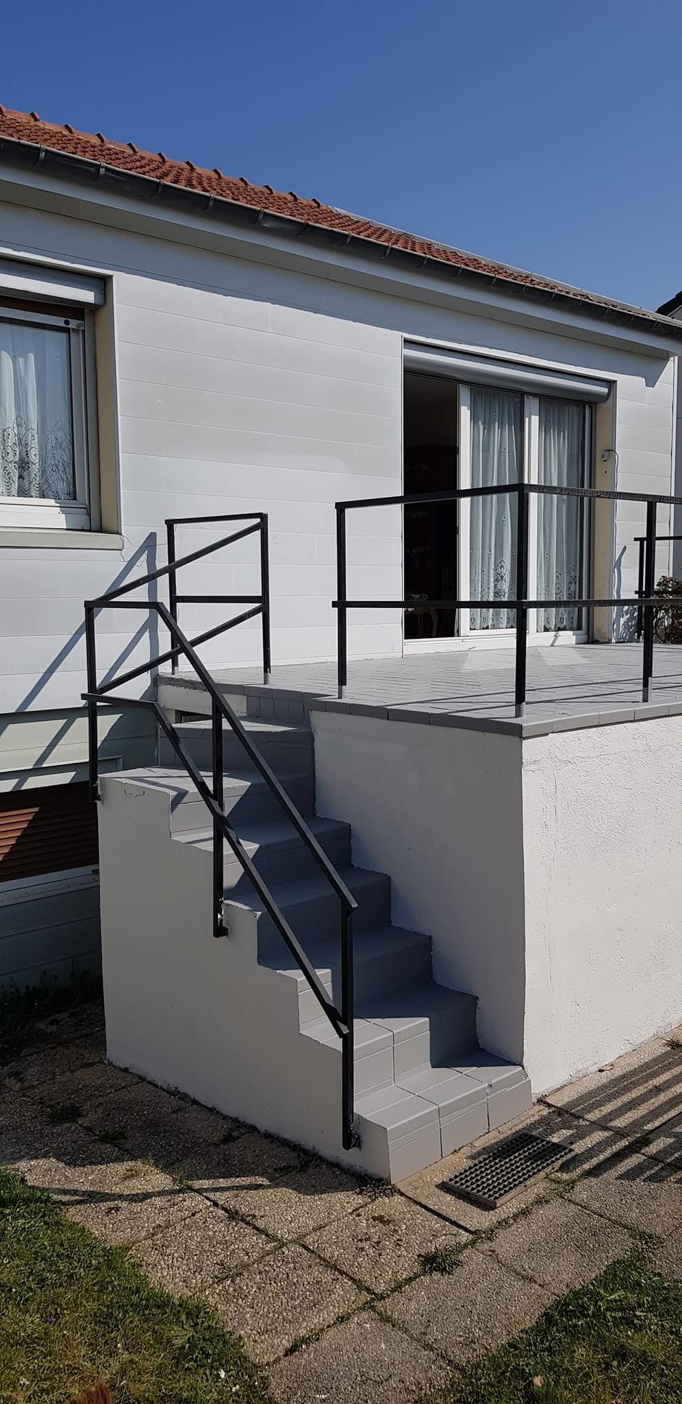 Petit escalier extérieur avec des rampes noires menant à l’entrée surélevée d’une maison blanche sous un toit de tuiles.
